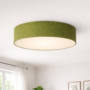 May include: A circular ceiling light with a green fabric shade. The light fitting has a white interior and a textured green exterior. The fixture is mounted on a white ceiling. The room has a window and artwork.