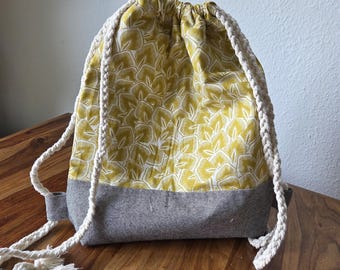 Könnte beinhalten: Ein Rucksack mit Kordelzug, der im oberen Bereich ein gelbes und weißes Blättermuster und im unteren Bereich einen grauen Stoff aufweist. Der Rucksack hat dicke, cremefarbene Seilriemen.