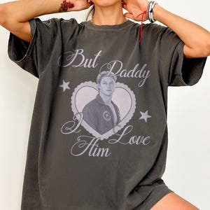 Könnte beinhalten: Ein anthrazitfarbenes Kurzarm-T-Shirt mit einem zentralen Aufdruck eines Mannes in einem herzförmigen Rahmen mit gewelltem Rand. Der Text "But Daddy I Love Him" ist in Schreibschrift gedruckt, ergänzt durch zwei kleine Sternsymbole.