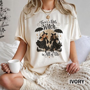 May include: An ivory short-sleeve t-shirt featuring a graphic print of two witches holding umbrellas under a full moon. The text reads, "There's a Little Witch in All of Us." The design includes silhouettes of pine trees and crescent moons.
