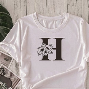 Peut inclure: T-shirt blanc avec une lettre H noire et un motif floral. La lettre H est centrée sur le t-shirt et le motif floral est à gauche de la lettre.