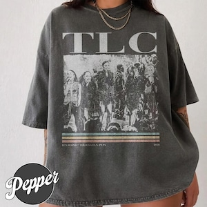 May include: A charcoal grey oversized t-shirt featuring a distressed graphic of the music group TLC. The print includes the text TLC, the phrase IT'S ICONIC TOUR SALT-N-PEPA 2026, and a series of horizontal coloured stripes below the group image.