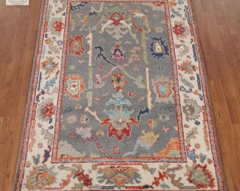 May include: A rectangular, hand-knotted rug with a gray central field and a cream-colored border. The rug features intricate floral and geometric patterns in shades of red, orange, blue, and green. The rug has a fringe on the short ends.