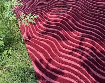 Może przedstawiać: Bordowy dywan z falistym wzorem w paski. Dywan jest prostokątny i wydaje się być wykonany z miękkiego materiału. Logo The Knotty Rug Co jest widoczne w lewym dolnym rogu. Dywan leży na zielonej trawie.
