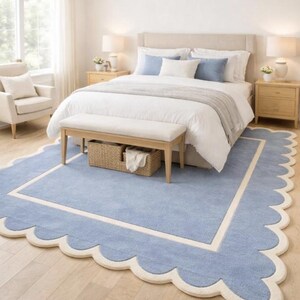 May include: A bedroom with a light blue scalloped-edge rug. The rug has a white border and is centered under a bed with white bedding and blue pillows. A bench and a wicker basket are at the foot of the bed.