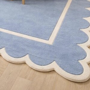 May include: A light blue rug with a scalloped edge and a cream-colored border. The rug has a soft, textured appearance and a rectangular design with a scalloped edge. The rug is placed on a light-colored wooden floor.