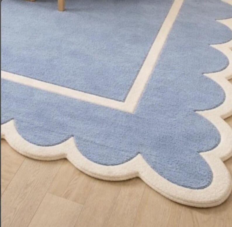 May include: A light blue rug with a scalloped edge and a cream-colored border. The rug has a soft, textured appearance and a rectangular design with a scalloped edge. The rug is placed on a light-colored wooden floor.