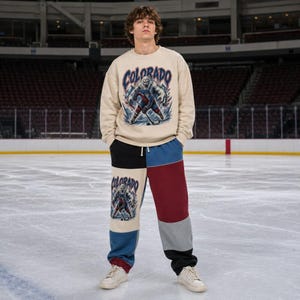 May include: A beige crewneck sweatshirt and color-blocked sweatpants featuring a graphic of a yeti hockey player and the text Colorado. The sweatpants are paneled in black, blue, maroon, and light gray, with the graphic on the left leg.