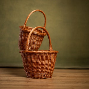 Può includere: Due cesti di vimini marroni con manici ad arco sono impilati. I cesti sono intrecciati e hanno un design classico, adatti per riporre oggetti o per l'esposizione. Lo sfondo è verde e la superficie è in legno.
