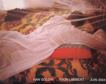 May include: A person sleeps under a white mosquito net on a bed with a patterned orange, yellow, and green bedspread. The person is wearing a white shirt and has a red pillow under their head. The text "NAN GOLDIN YVON LAMBERT JUIN 2004" is printed at the bottom of the image.