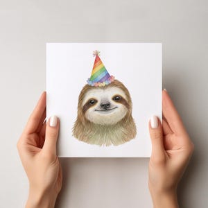 May include: A square greeting card showing a smiling sloth wearing a rainbow party hat. The sloth has brown fur and dark eyes. The party hat is cone-shaped with rainbow stripes and a pom-pom on top. The card is held by two hands.