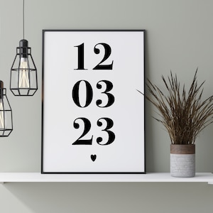 May include: Black and white print featuring the numbers 12, 03, and 23, with a small heart symbol at the bottom. The print is framed in a black frame and is hanging on a white shelf with a potted plant.