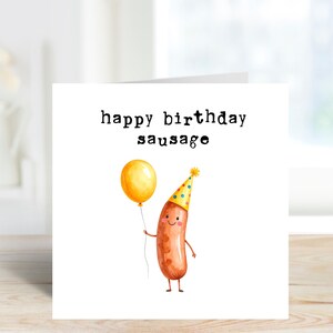 May include: A white greeting card with the text "happy birthday sausage". The card features a watercolor illustration of a smiling sausage wearing a party hat and holding a yellow balloon. The background is a blurred interior.