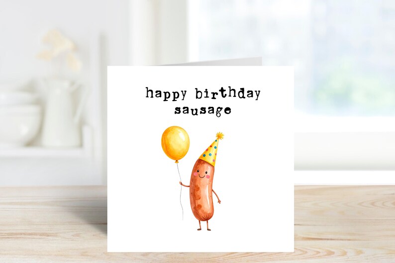 May include: A white greeting card with the text "happy birthday sausage". The card features a watercolor illustration of a smiling sausage wearing a party hat and holding a yellow balloon. The background is a blurred interior.