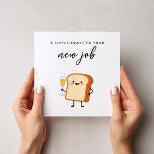May include: A white card with the text "A LITTLE TOAST TO YOUR new job" in black script. The card features a cartoon illustration of a slice of toast holding a champagne glass. The card is held by two hands.