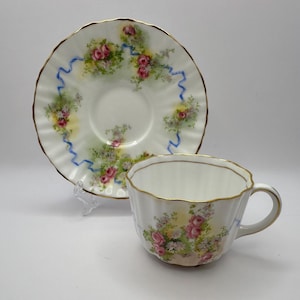 Könnte beinhalten: Ein weißes Tee-Service mit floralem Design. Die Tasse und Untertasse haben einen Goldrand und einen gewellten Rand. Das florale Design umfasst rosa Rosen, grüne Blätter und blaue Bänder. Das Set wird auf einer weißen Oberfläche präsentiert.