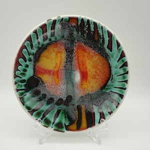 May include: A circular ceramic plate with a white edge, showcasing a vibrant abstract pattern. The design incorporates orange, yellow, green, and black hues. The plate is presented on a clear plastic stand, highlighting its decorative purpose.