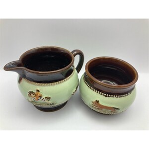 May include: A ceramic creamer and sugar bowl set. The creamer has a spout and handle, while the sugar bowl is round. Both pieces are light green with brown rims and bases. The creamer has a floral design, and the sugar bowl has a fox.