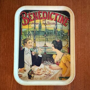 以下が含まれることがあります： ベネディクティンリキュールのヴィンテージ広告が描かれた長方形のサービングトレイ。トレイには、ボトルとペストリーのあるテーブルにいるカップルが描かれており、ベネディクティンのロゴとステンドグラスの背景があります。