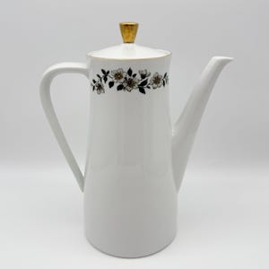 May include: White ceramic coffee pot with a gold-coloured lid and handle. The pot features a floral design in black and white around the top. The spout is long and slender, and the handle is curved.
