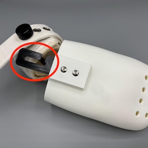May include: A white protective boot with a black plastic hook and a cream-colored strap. The boot has several small holes and is secured with two silver screws. A black knob is attached to the strap.