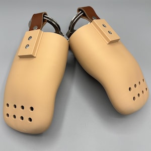 May include: Two beige shoe trees with brown leather straps and metal rings. Each tree has a rectangular accent piece and several small holes near the toe. The shoe trees are designed to maintain the shape of shoes.