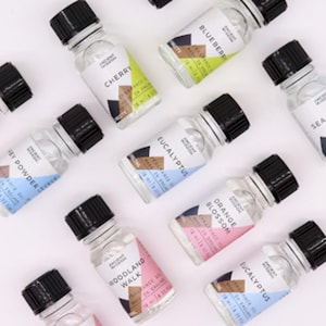 May include: A collection of small glass bottles with black lids, each displaying a different fragrance name. Labels include "Cherry," "Blueberry," and "Eucalyptus," alongside the brand name "Ancient Wisdom." Each bottle contains 10 ml / 0.33 fl oz.
