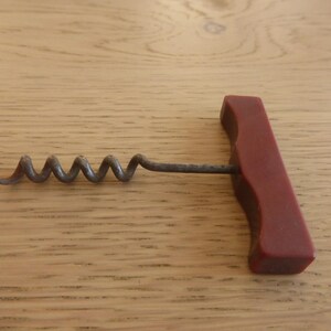 May include: A vintage corkscrew with a dark red handle and a metal worm. The corkscrew is designed for removing corks from wine bottles. The handle is rectangular and has a smooth finish.