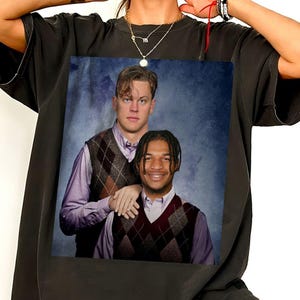 May include: A black t-shirt featuring a graphic print of two individuals posing for a portrait. Both wear light-colored collared shirts under argyle sweater vests. The background of the print is a mottled blue texture. The shirt has short sleeves.