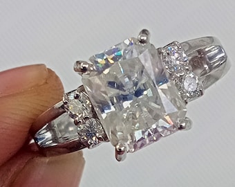 Puede incluir: Un anillo de plata con una gran piedra preciosa rectangular transparente rodeada de piedras preciosas transparentes más pequeñas.