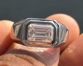 May include: A silver-toned ring featuring a rectangular emerald-cut clear stone set in a smooth, polished metal bezel. The band has a wide, solid construction that tapers slightly toward the back, showcasing a clean, reflective metallic finish.