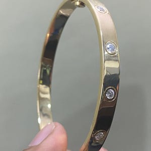 May include: A polished gold bangle bracelet featuring a series of evenly spaced, round, clear, faceted stones set into the metal band. The bracelet includes a visible hinged clasp mechanism at the top, showcasing a smooth, reflective metallic finish.