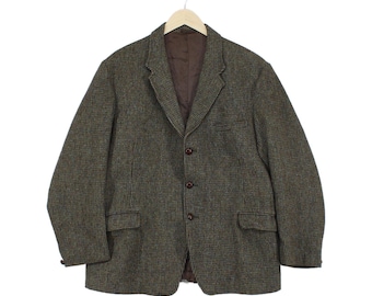 Pode incluir: Um blazer de tweed castanho e verde-azeitona com um padrão pied-de-poule texturado, lapela entalhada, três botões castanhos escuros no fecho frontal, dois bolsos com aba na cintura e forro interior castanho escuro liso num cabide.