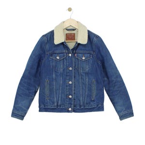 Puede incluir: Una chaqueta de mezclilla azul con cuello de sherpa color crema, cierre frontal de botones metálicos, dos bolsillos en el pecho con solapas abotonadas y dos bolsillos laterales. Una etiqueta de marca tipo cuero marrón es visible en el cuello.