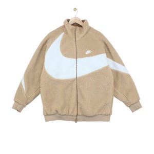 Op de afbeelding: Een beige, pluizig fleecejack in sherpa-stijl met een volledige ritssluiting aan de voorzijde en een hoge kraag. Een groot, wit, gebogen Nike swoosh-logo loopt over de borst en mouwen. Een klein wit Nike-logo is op de linkerborst geborduurd.