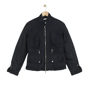 May include: A black jacket featuring a high collar, a full-length silver-tone front zip, and long sleeves with zipped cuffs. The garment includes two horizontal zipped chest pockets and two lower flap pockets with silver-tone snap buttons.