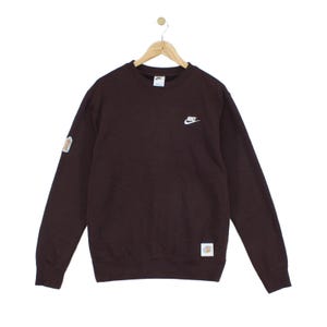 May include: A dark brown long-sleeve crewneck sweatshirt featuring a small white Nike logo embroidered on the upper left chest. The garment includes a rectangular patch on the lower left hem and a smaller patch on the right sleeve near the shoulder.