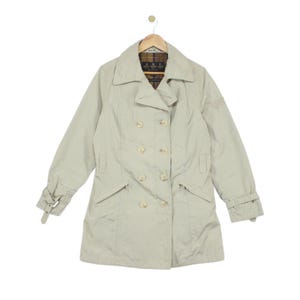 May include: A light beige double-breasted trench coat featuring a wide collar, long sleeves with adjustable buckled cuffs, and two slanted front pockets. The interior lining displays a dark brown, yellow, and black plaid pattern with a Barbour label.