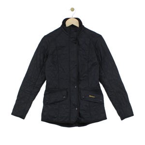 May include: A black quilted jacket featuring a high collar, long sleeves, and a front snap-button closure. The garment includes two front flap pockets with snap buttons and the word Barbour embroidered in gold thread on the right pocket.