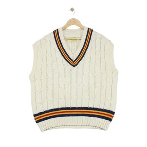 May include: An off-white cable knit sweater vest featuring a V-neckline and a ribbed hem. The neckline and hem are accented with horizontal stripes in navy blue, red, and yellow. The garment is displayed on a wooden hanger against a plain background.
