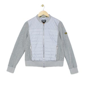 May include: A light grey bomber jacket featuring a quilted front panel, long sleeves, and a ribbed collar, cuffs, and hem. The jacket includes a full-length silver front zip, two zipped side pockets, and a Barbour brand label at the neck.