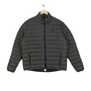 May include: An olive green puffer jacket with a high collar, long sleeves, and a full-length black front zip. The jacket features horizontal quilted stitching, two zipped side pockets, and the black letters CK embroidered on the chest.