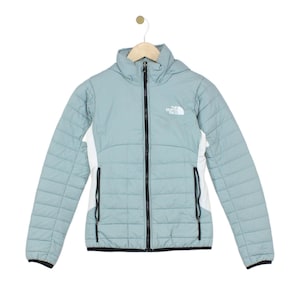May include: A light blue, quilted, hooded jacket by The North Face featuring a full-length black front zip, two black zipped side pockets, and white side panels. The brand logo is embroidered in white on the upper left chest area.