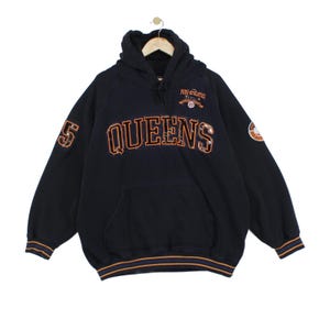 May include: A navy blue hooded sweatshirt featuring the word QUEENS in large orange and white outlined lettering across the chest. The garment includes a front kangaroo pocket, ribbed cuffs with orange and blue stripes, and FUBU ATHLETICS text.