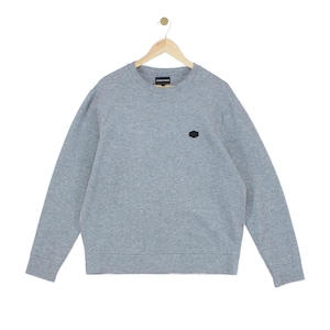 May include: A heather gray long-sleeve crewneck sweatshirt featuring a small black hexagonal patch on the left chest and a dark rectangular brand label inside the neckline. The garment has ribbed cuffs and a ribbed hem, hanging on a wooden hanger.