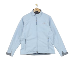 Könnte beinhalten: Eine hellblaue Softshelljacke mit durchgehendem Frontreißverschluss, hohem Kragen und zwei seitlichen Reißverschlusstaschen. Das The North Face Logo ist auf der linken Brust gestickt. Die Jacke hat lange Ärmel mit verstellbaren Bündchen.