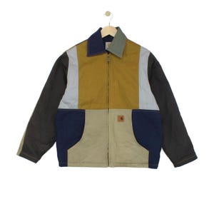 Könnte beinhalten: Eine farblich abgesetzte Reißverschlussjacke mit einem senfgelben Mittelteil, hellgrauen Seitenteilen, dunkelbraunen Ärmeln und marineblauen unteren Taschen. Der Kragen ist in Marineblau und Salbeigrün geteilt, mit einem kleinen Carhartt-Logo-Patch.