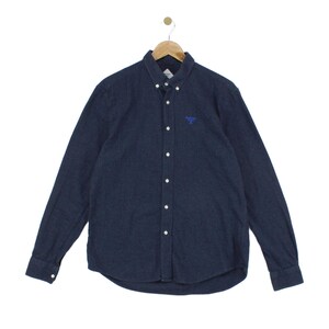 Puede incluir: Una camisa de manga larga con botones en un tejido texturizado de color azul marino oscuro. La camisa presenta un cuello clásico, una tapeta frontal con botones blancos y un pequeño logotipo bordado en azul en el lado izquierdo del pecho. Se muestra en una percha.