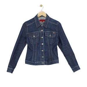 May include: A dark blue denim jacket featuring a classic collar, long sleeves with buttoned cuffs, and a front button closure. The jacket includes two chest pockets with buttoned flaps, vertical seam detailing, and a red interior neck label.