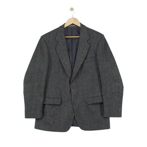 May include: A dark gray blazer featuring a subtle herringbone pattern, a notched lapel, two front buttons, and two flap pockets. The jacket has long sleeves with three buttons on each cuff and is displayed on a wooden hanger against a white background.